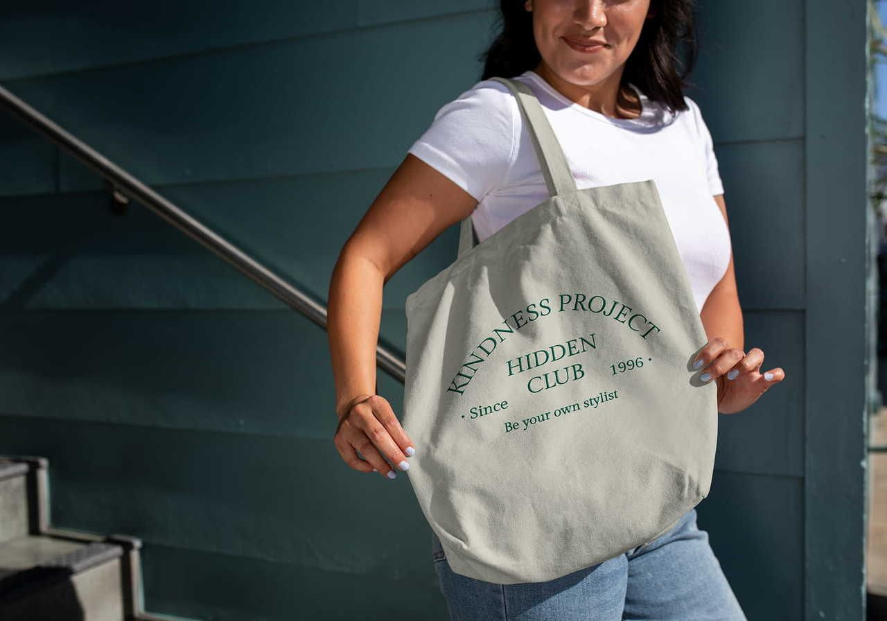 A woman showing a custom tote bag with “Kindness project” and “Hidden club” text prints. Blue wooden wall behind her.