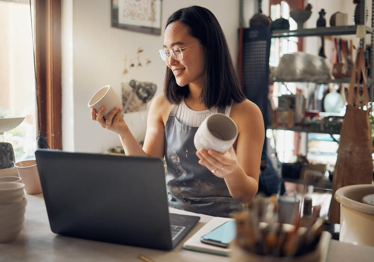 A young woman artist is holding two mugs while sitting at a desk in front of a laptop.