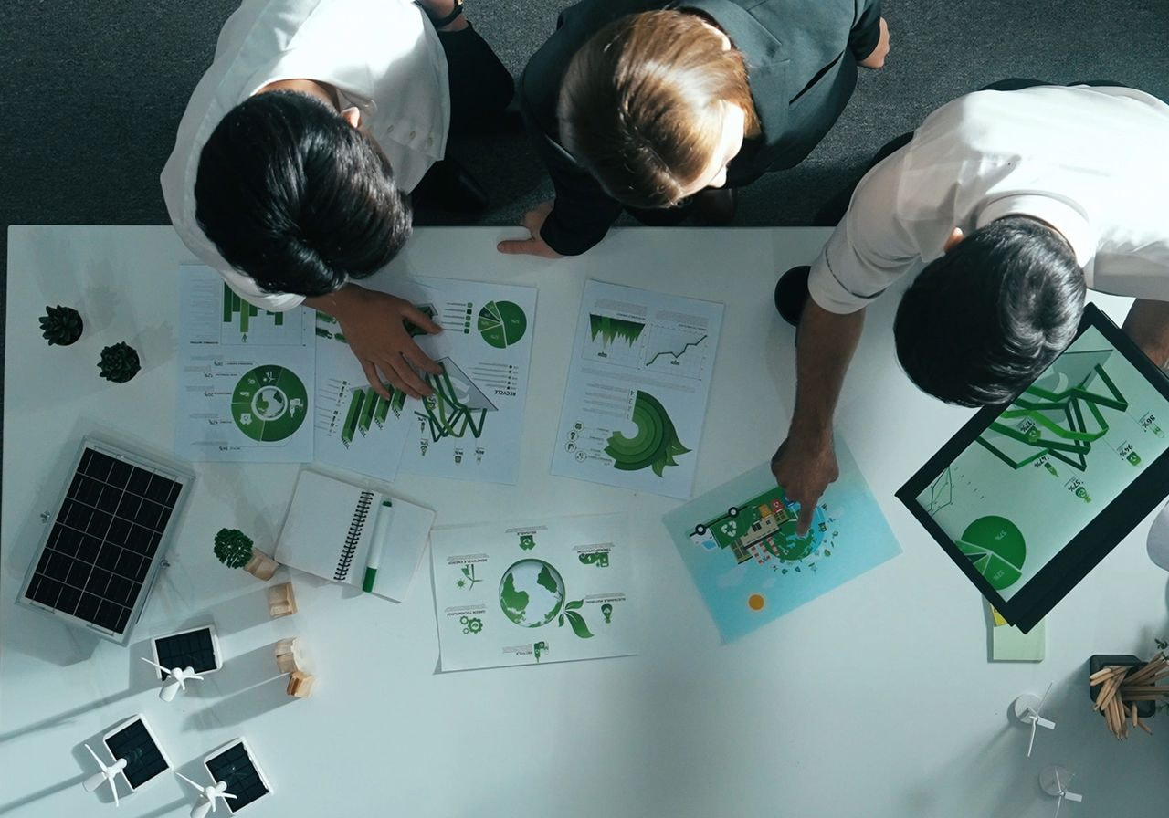 A team reviews green sustainability charts and infographics on a white table with model solar panels and wind turbines, seen from above.