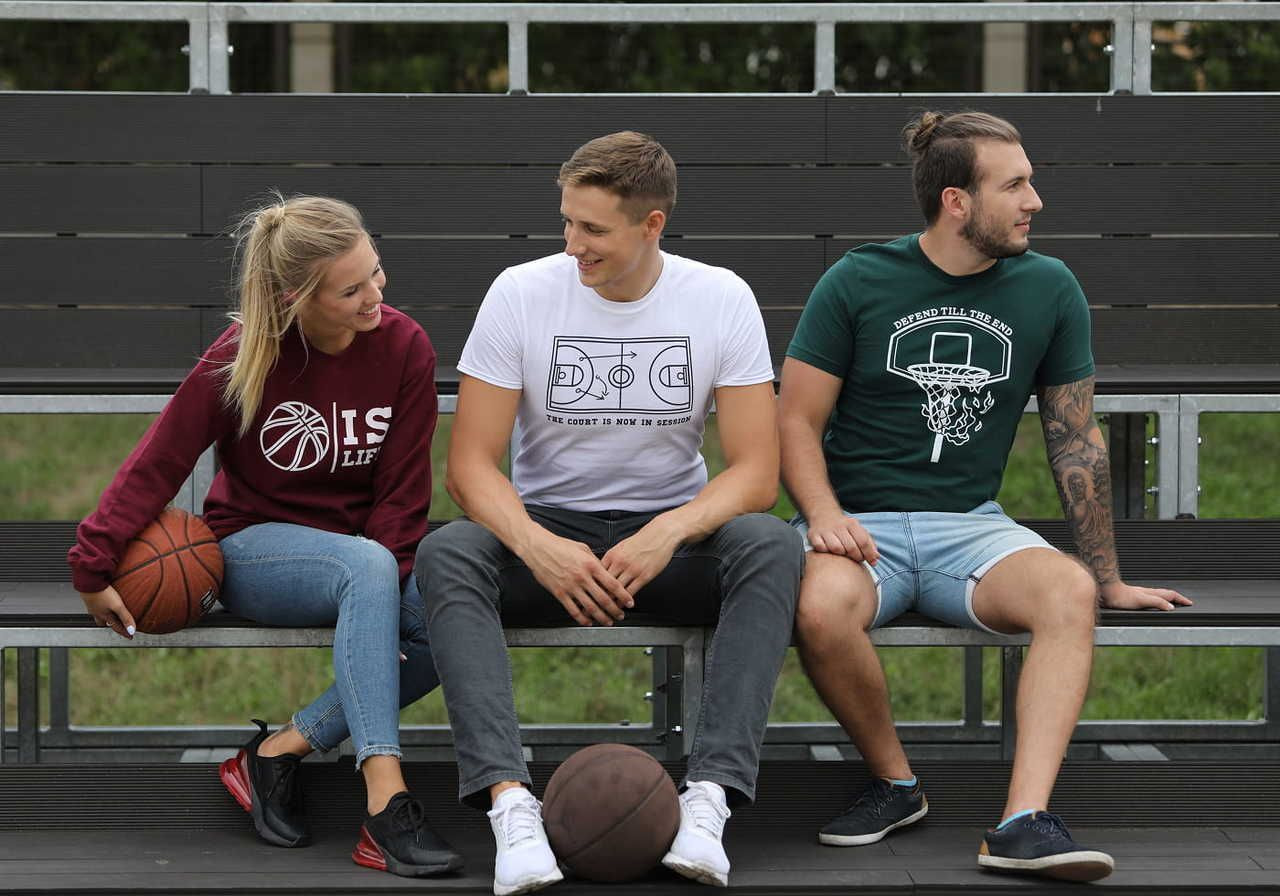 Three adults on bleachers: a woman in a maroon sweatshirt with a basketball, a man in a white t-shirt, and another man in a green t-shirt.