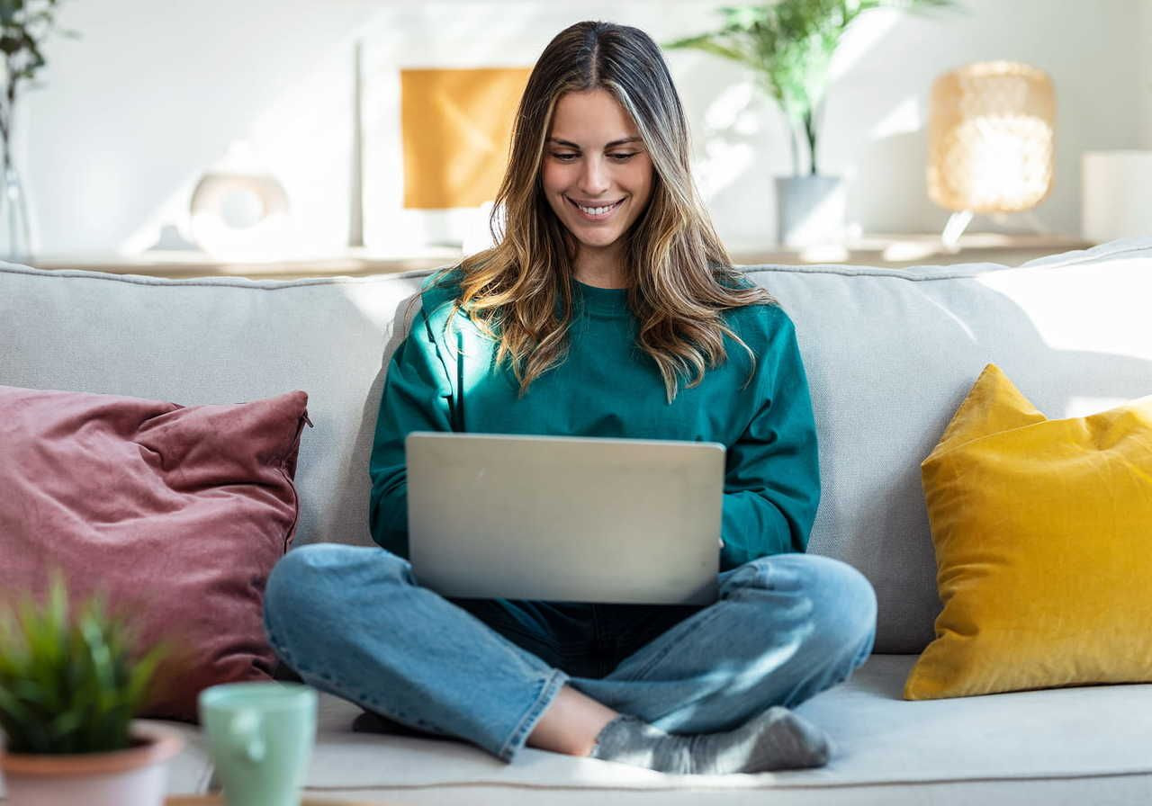 A smiling woman in a green sweater sits cross-legged on a sofa, using a laptop to learn how to make custom patches using Printful.