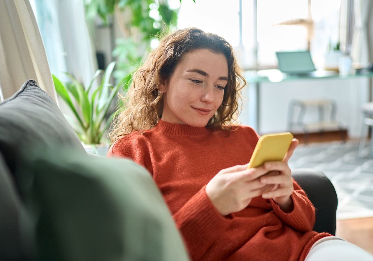 Woman with curly hair smiling while using her phone on a couch.