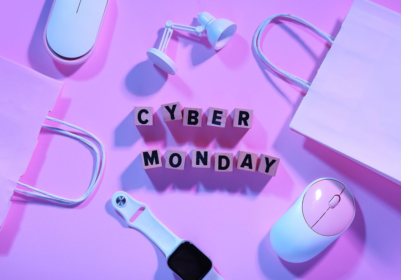 Wooden letter blocks spelling “Cyber Monday” surrounded by shopping bags, computer mice, a smartwatch, and a mini desk lamp on a pink background.