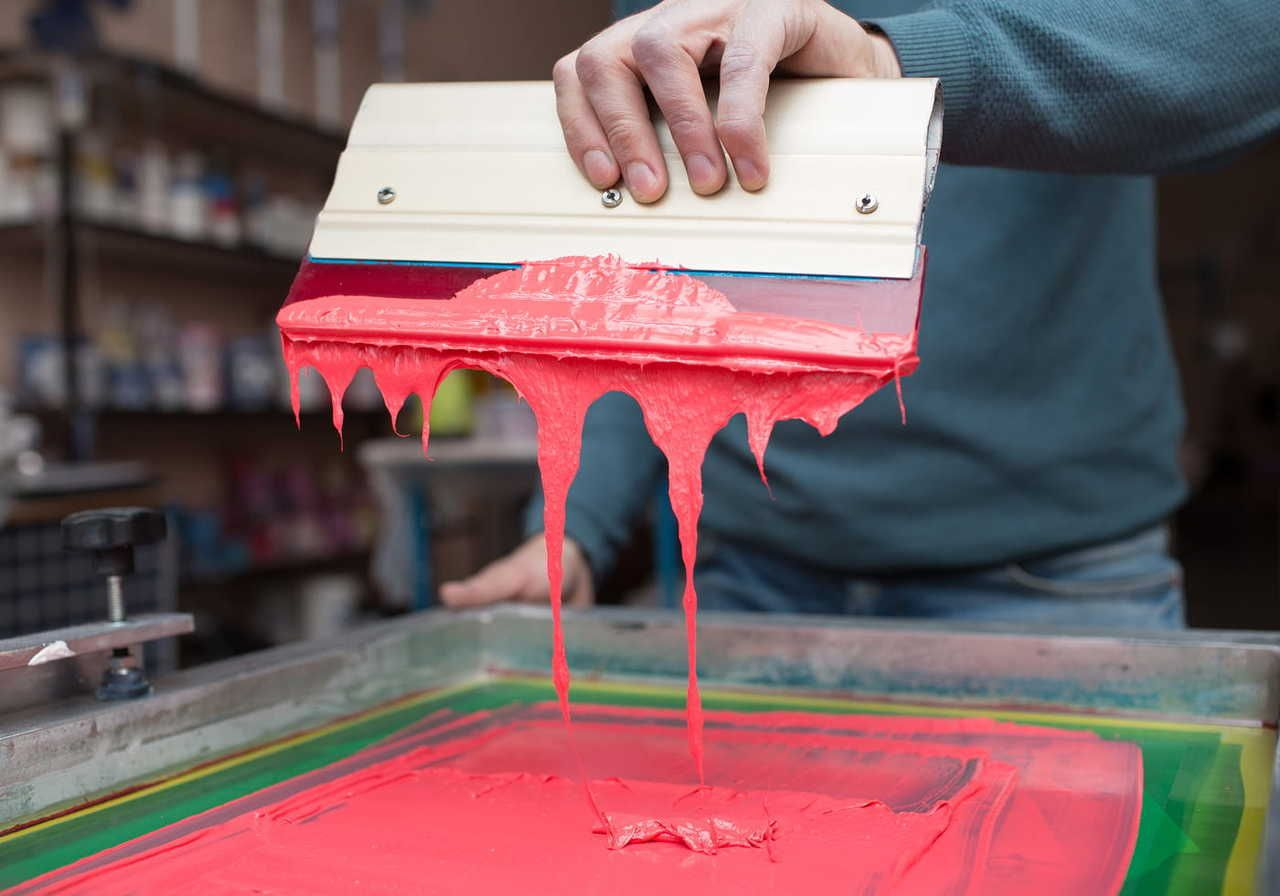 A person using a squeegee to spread bright pink paint over a t-shirt on a screen printing machine.