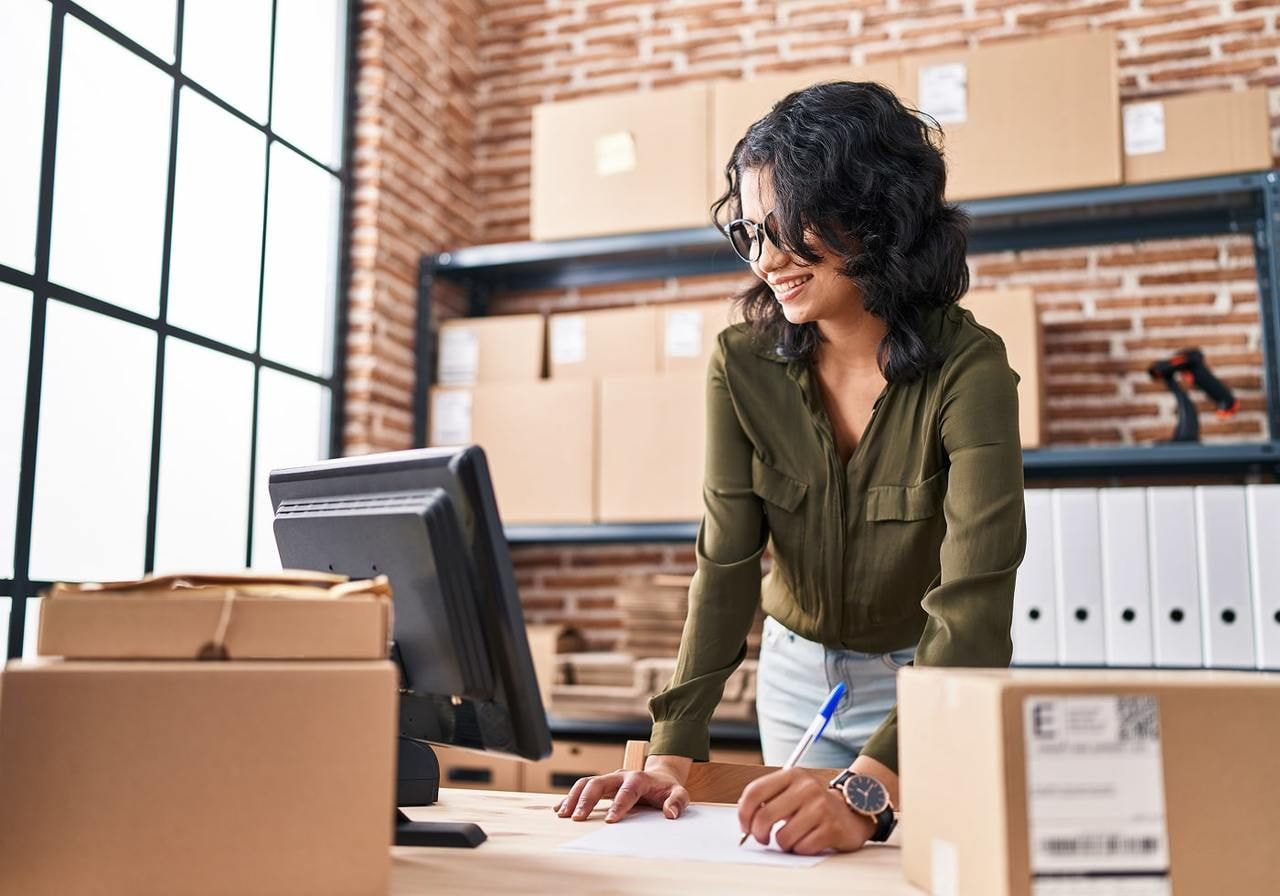 A woman wearing glasses and a green blouse smiles while writing on a paper in a office, with packaged in the background.