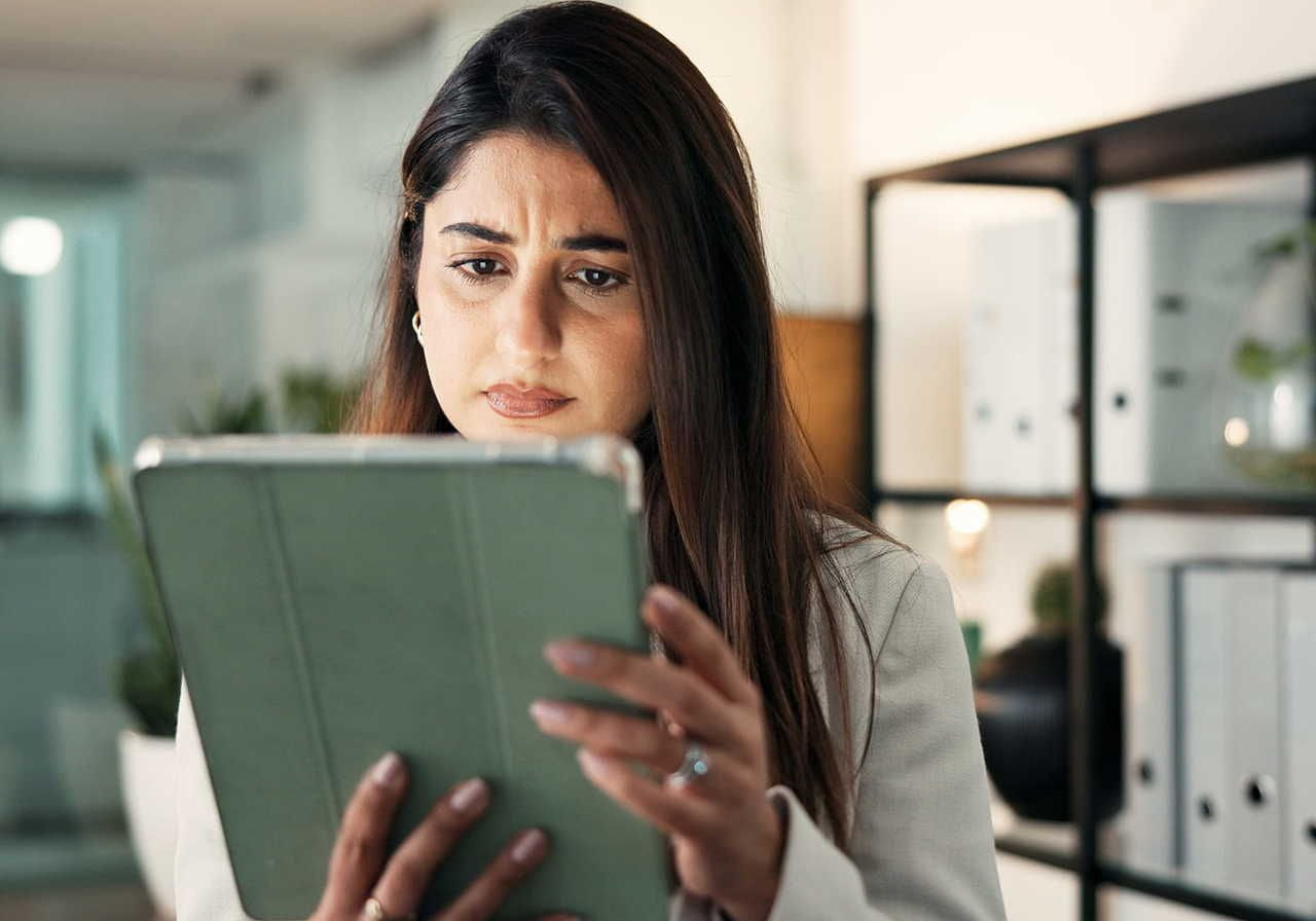 A woman with long dark hair looks concerned while holding a tablet in a bright office. Behind her are shelves with books and plants.