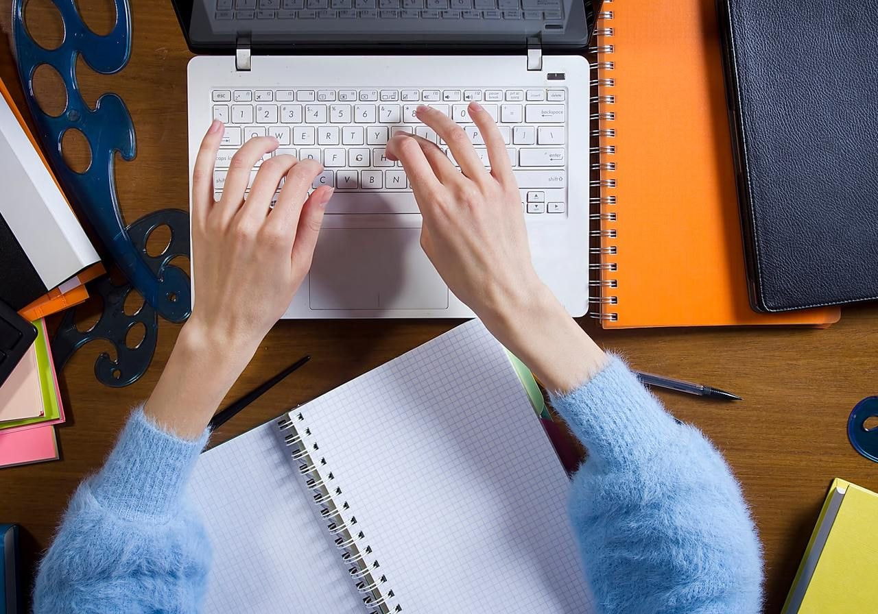 Hands typing on a white laptop keyboard, surrounded by notebooks, papers, and drafting tools on a wooden desk.