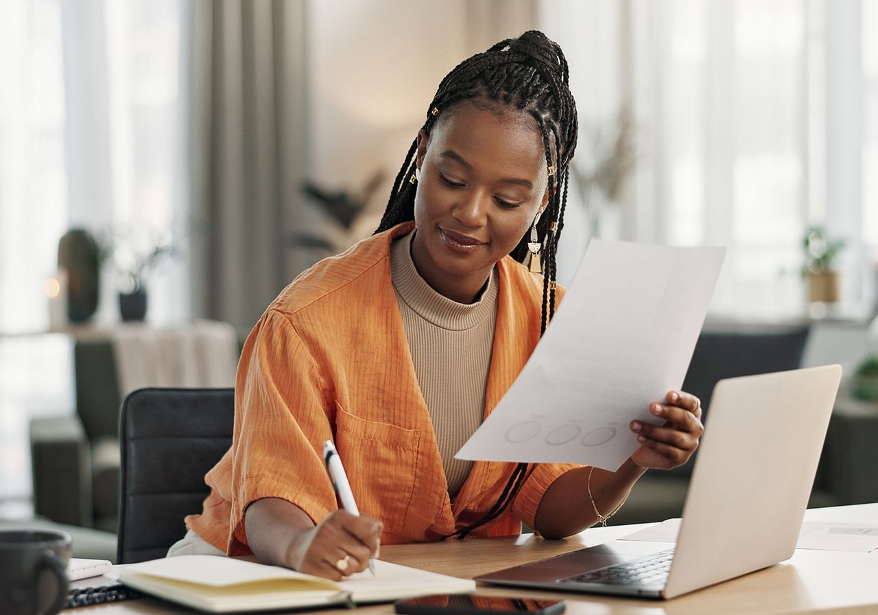 A woman in an orange jacket writes notes at a desk, holding a document. 