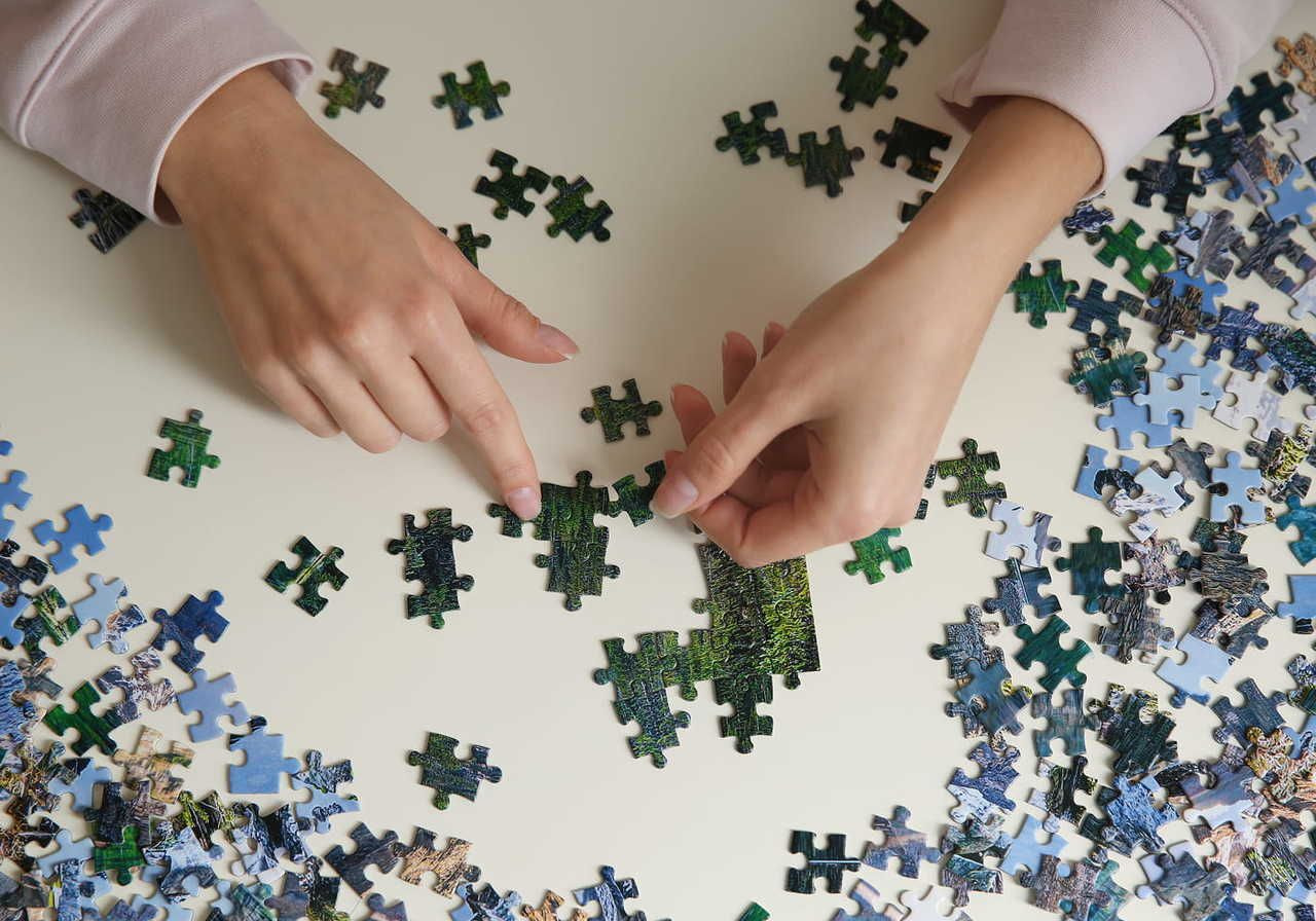 Hands arranging jigsaw puzzle pieces on a white table, surrounded by scattered pieces.