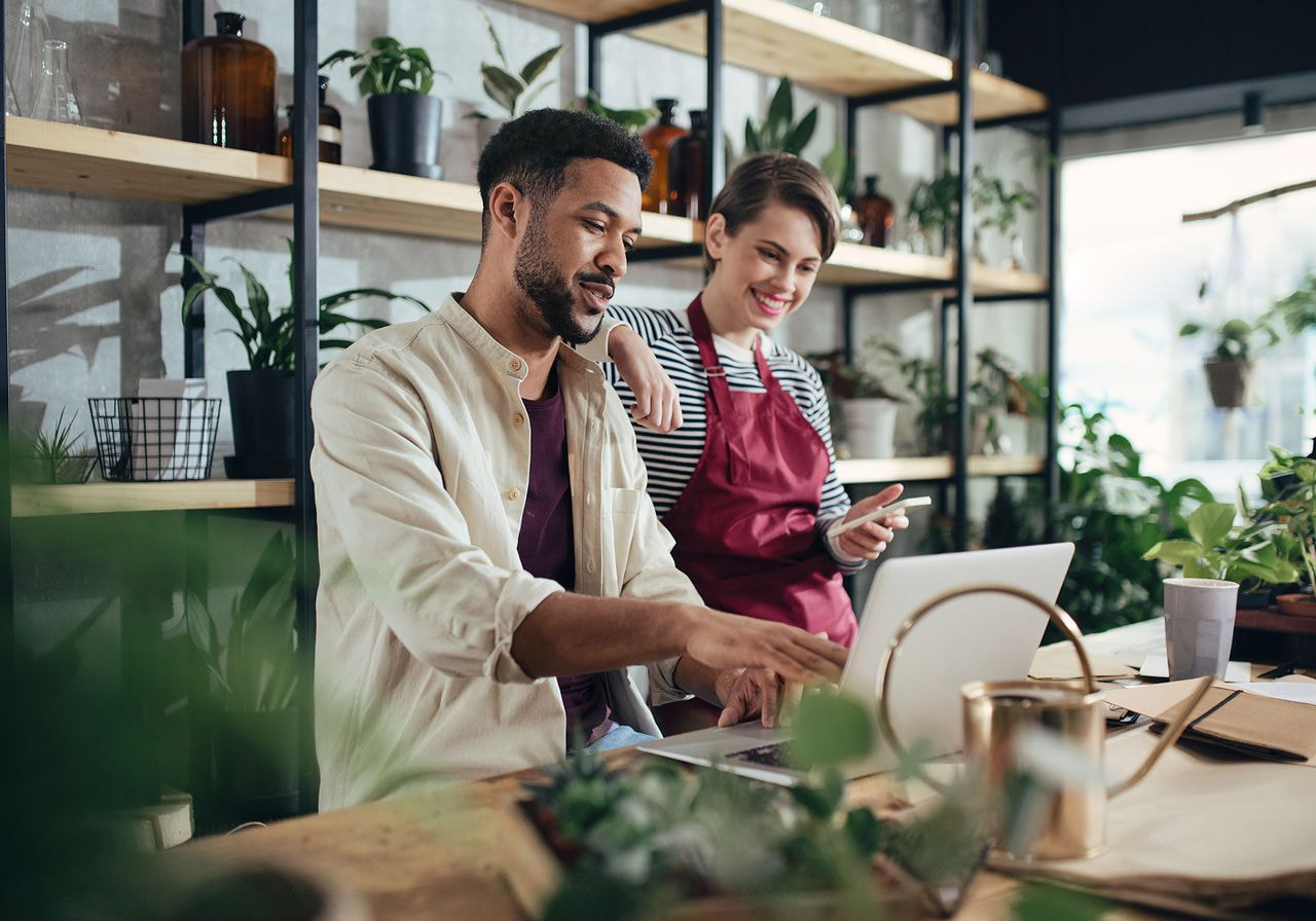 A man and woman collaborate on a laptop amidst colorful flowers in a vibrant flower shop.