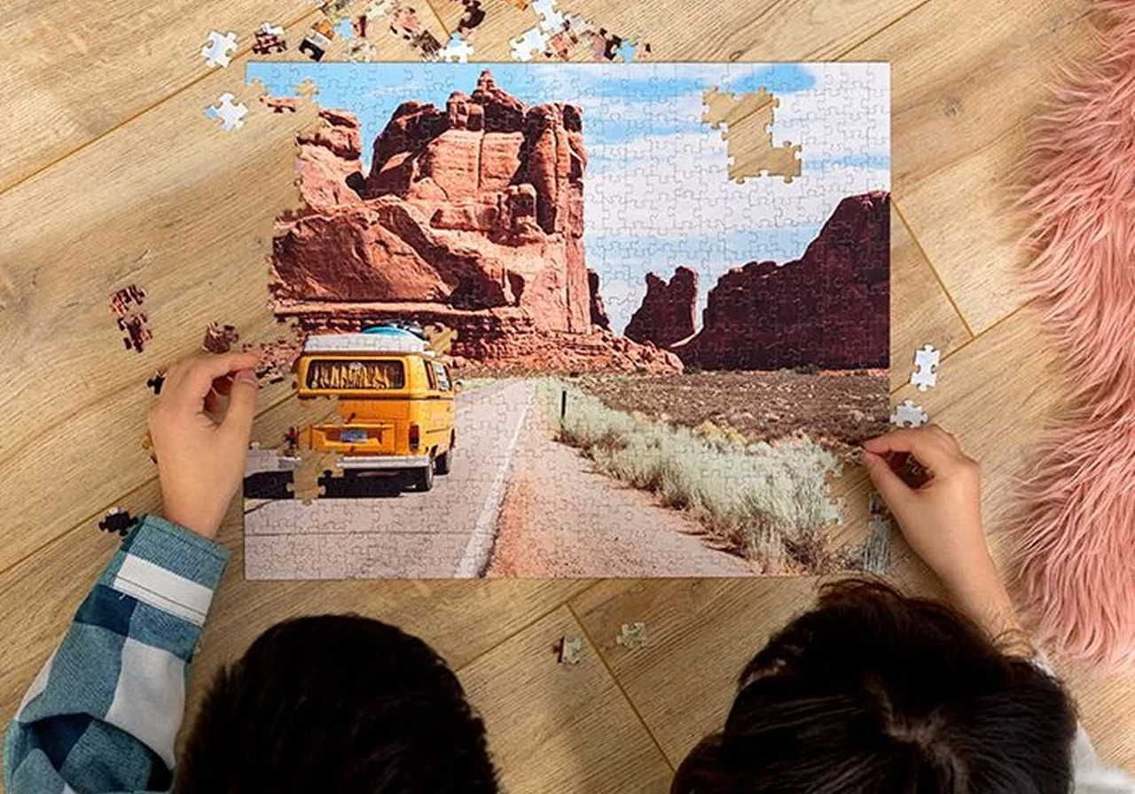 Two people are assembling a puzzle showing a yellow van on a desert road with red rock formations.