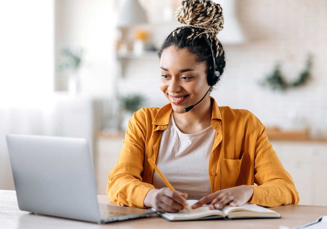 A smiling woman wearing headphones with a mic, is looking at a laptop and writing in a notebook.