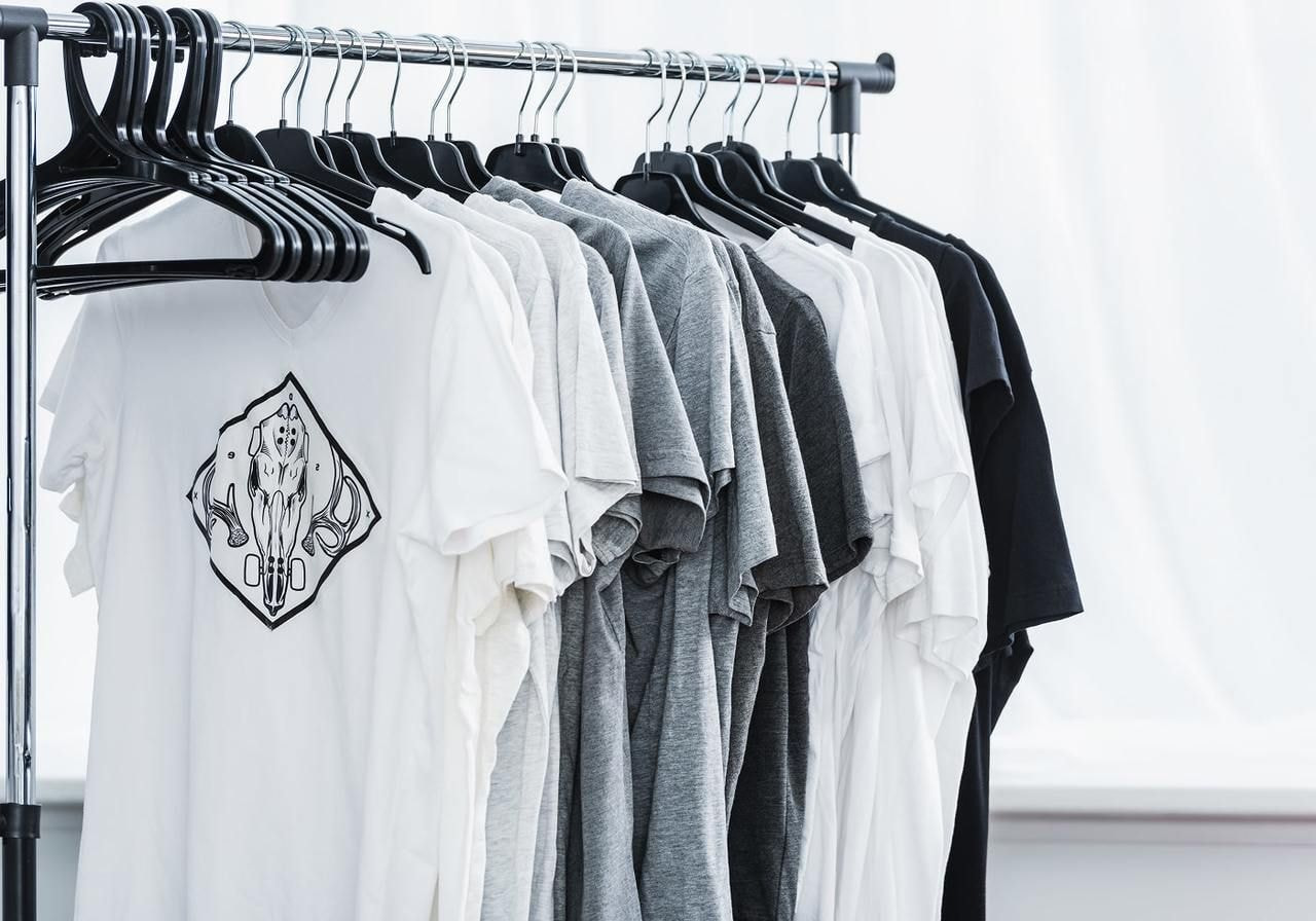 A row of white, gray, and black t-shirts on hangers, hanging on a rack. One white shirt features a geometric design.