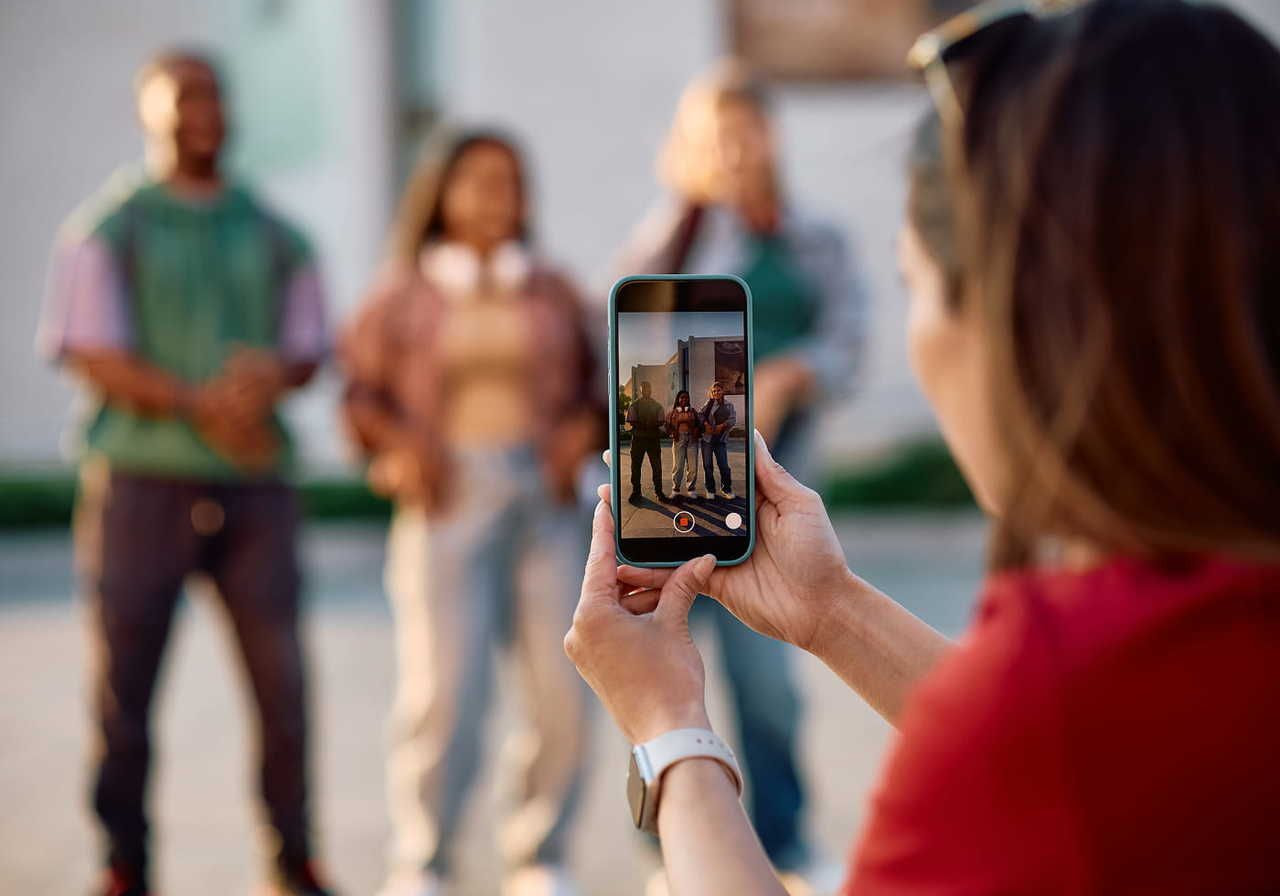 A person in the foreground uses a smartphone to capture three smiling friends posing outdoors.