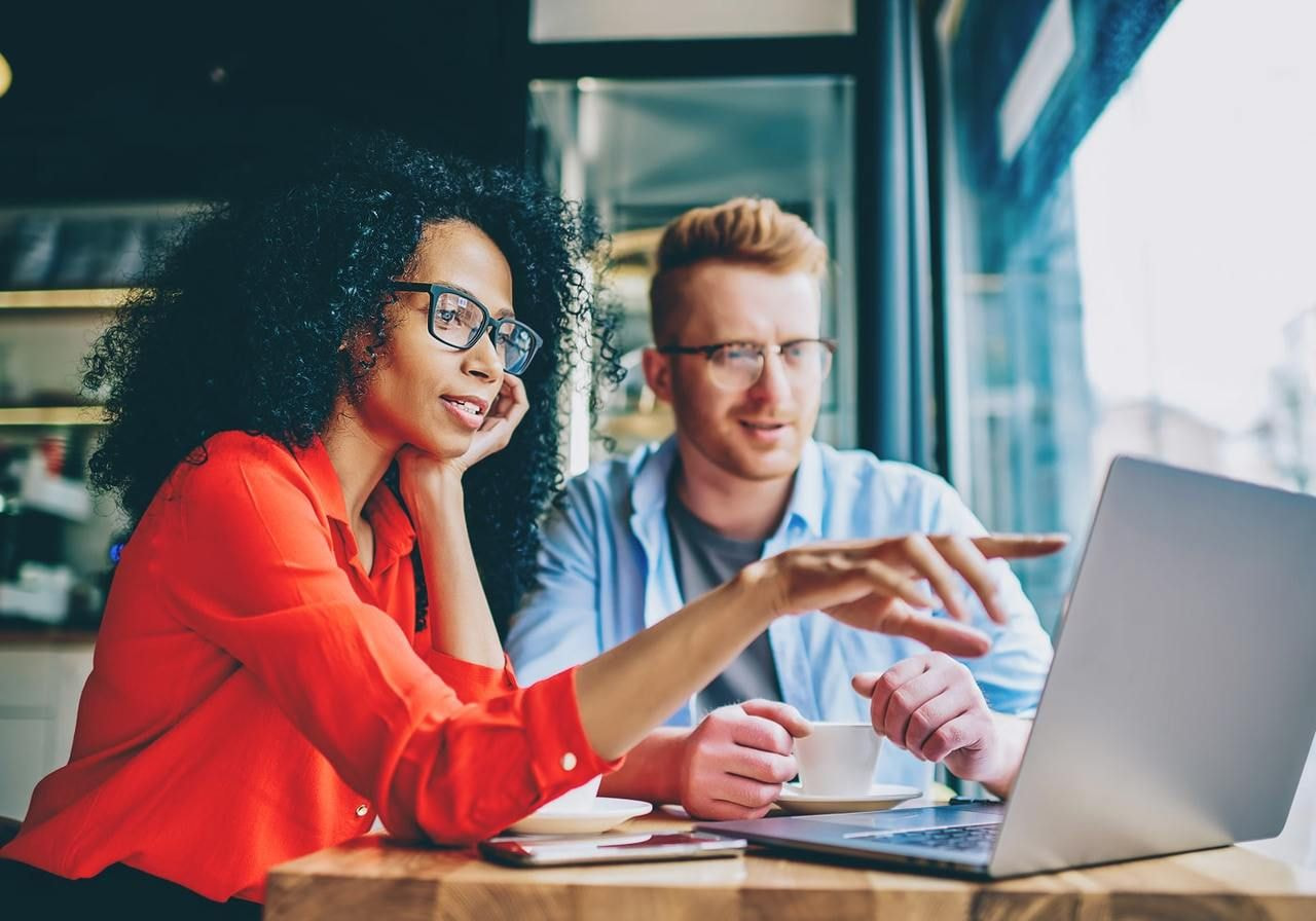 A woman in a red shirt and a man in a blue shirt discuss starting a hoodie business. The woman points at a laptop screen.