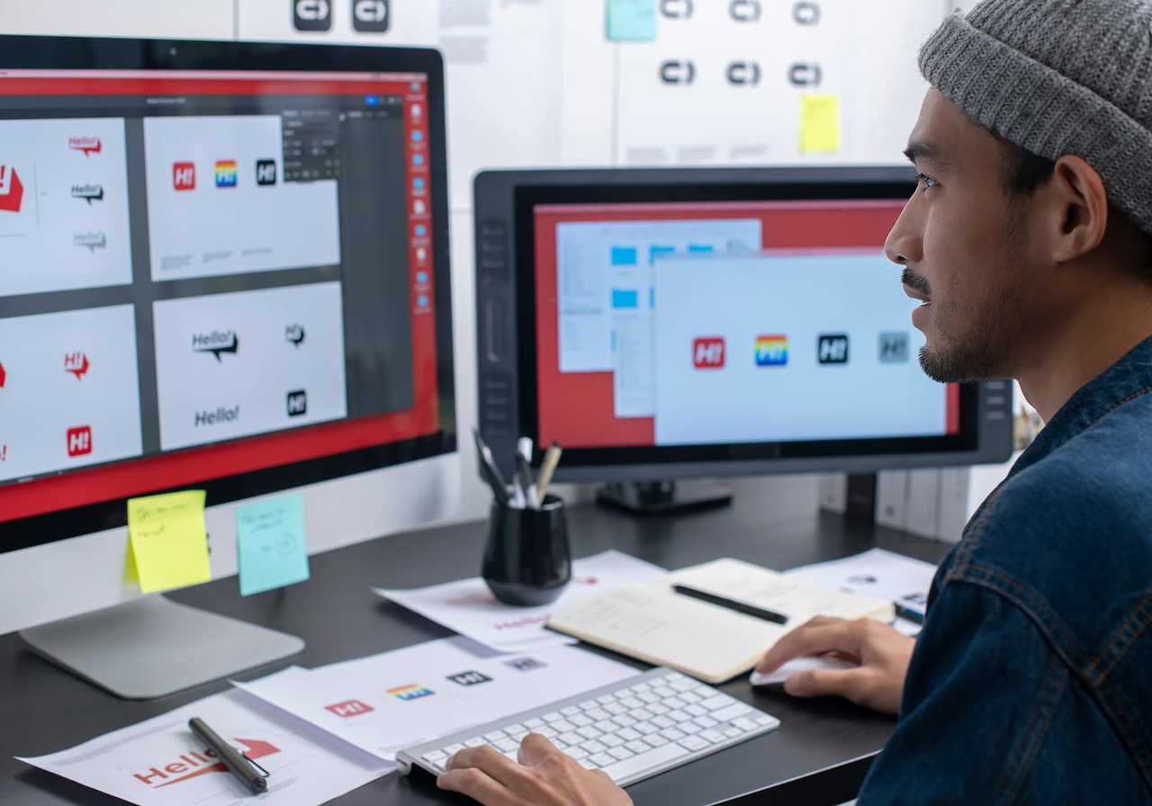 A man in a beanie focuses on a computer with multiple logo designs. The workspace is organized with notes and stationery.