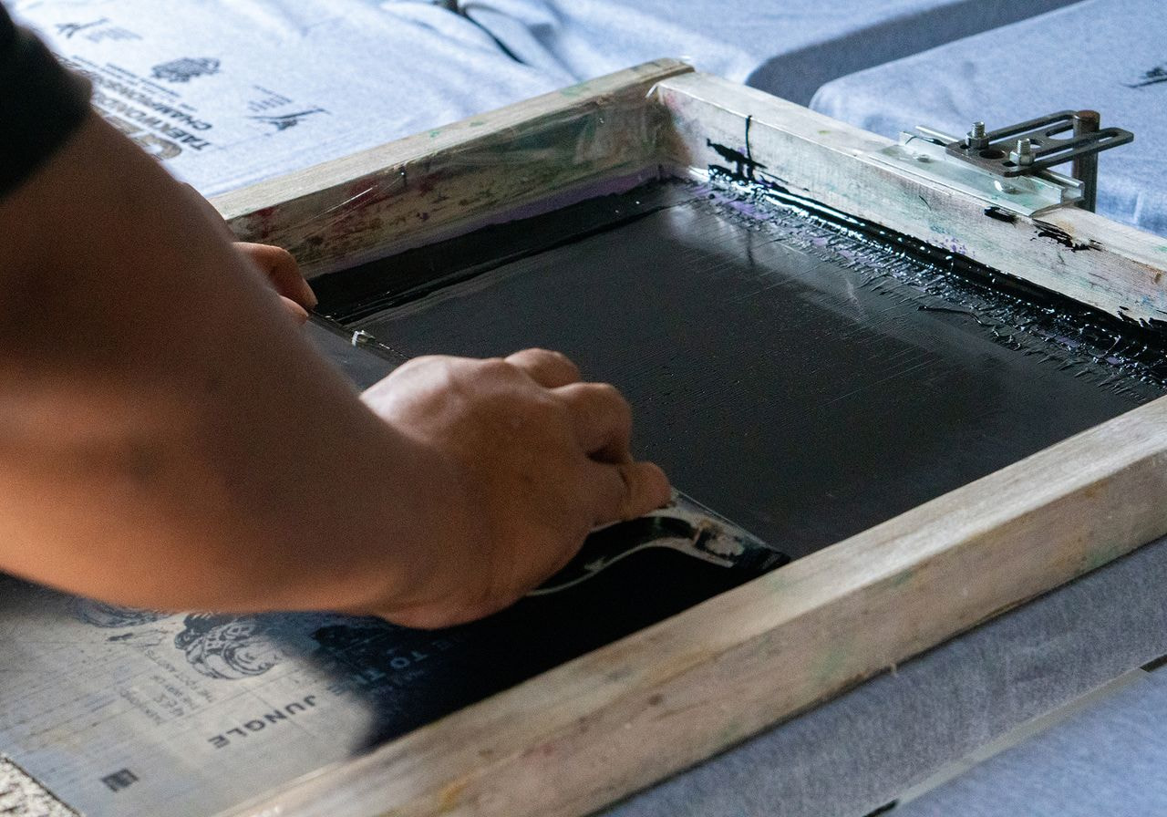 Close-up of a hand using a squeegee to screen print black ink onto gray fabric.