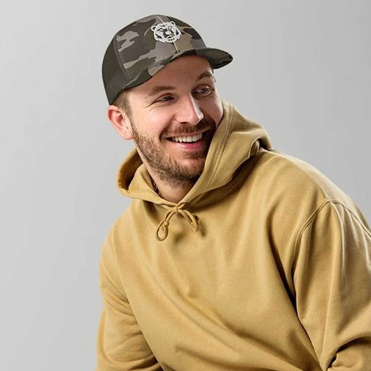 A smiling man in a tan hoodie and camo baseball cap with a white embroidered bear logo, set against a plain gray background.