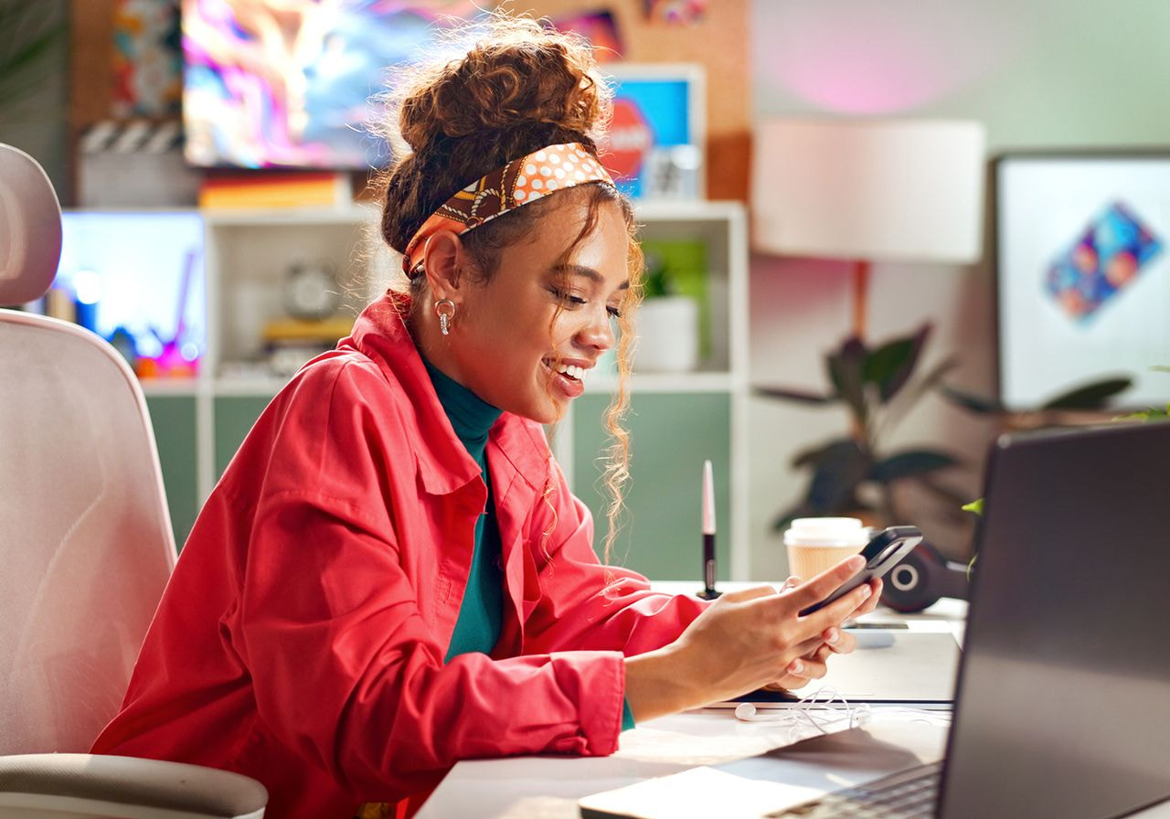 A woman in a red jacket and headband sits at a desk, smiling at her phone in a colorful, cozy workspace with plants and a laptop.