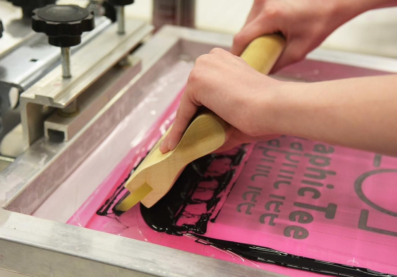 Hands use a squeegee to spread black ink over a pink screen with text, in a screen printing process.