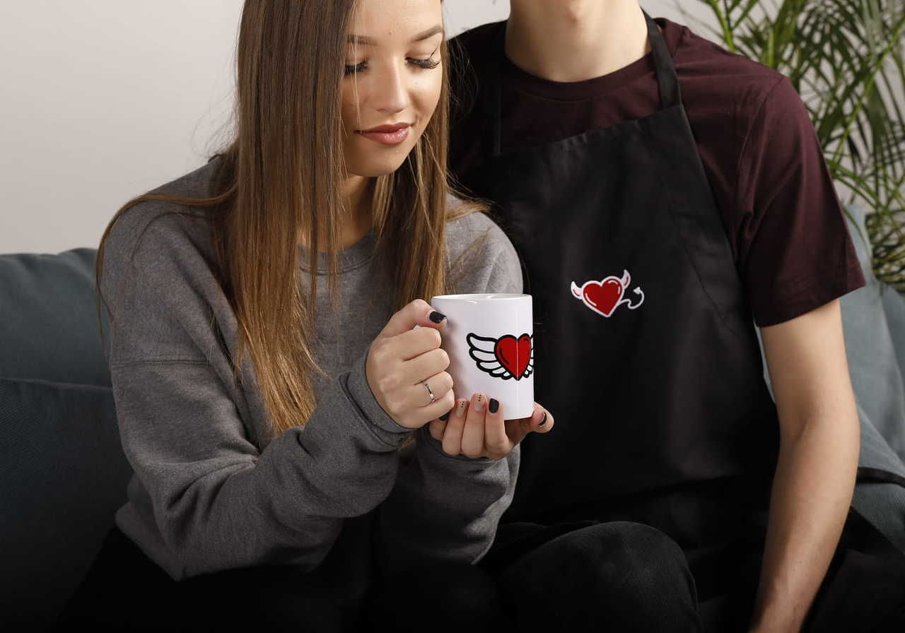 A young woman sitting on a couch holding a mug with a heart and wings design, while a young man in an apron sits beside her.