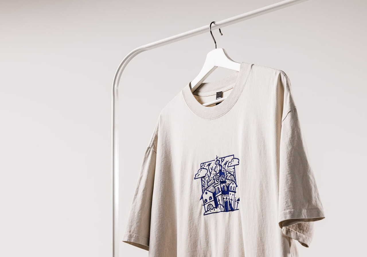 A light beige t-shirt with a blue graphic design featuring a house and clouds hanging on a hanger against a neutral background.