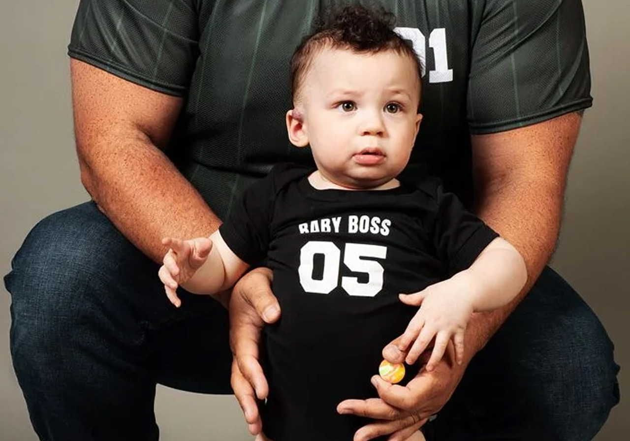 A baby wearing a black "Baby Boss 05" shirt stands in front of an adult. The adult gently supports the baby with both hands.