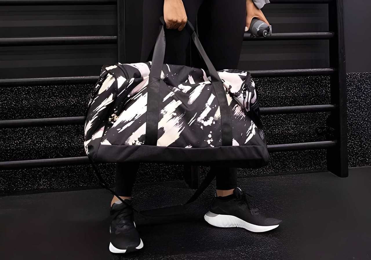 Person holding a stylish, black and white patterned gym bag, wearing black athletic shoes and leggings, standing in a gym setting.
