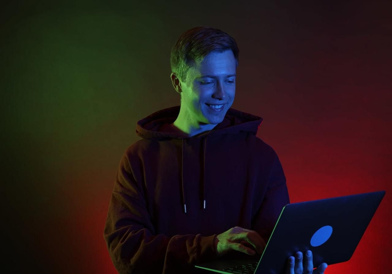 A person wearing a hoodie smiles while using a laptop, illuminated by blue, green, and red lights.