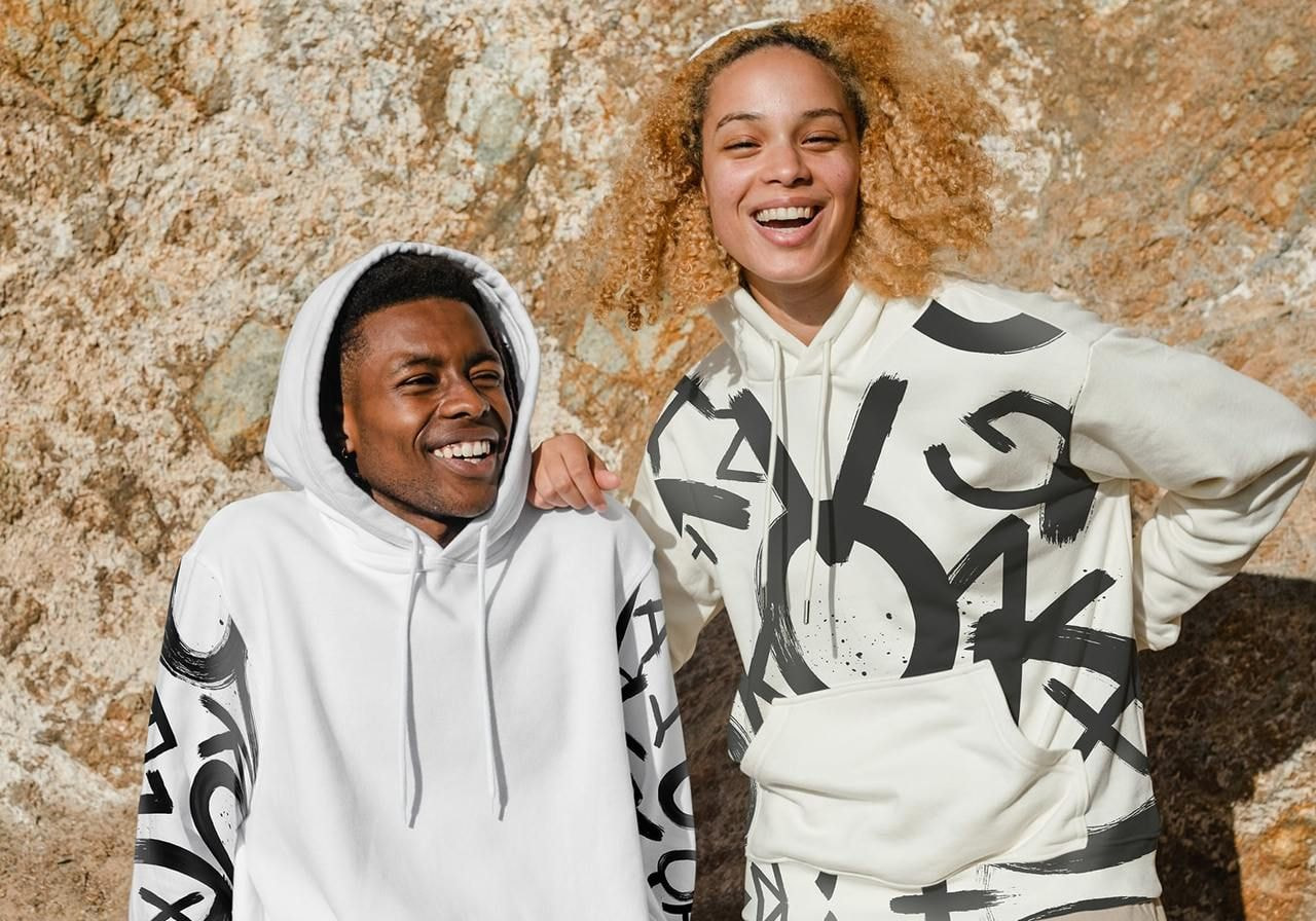 Two people smiling and wearing white hoodies with bold, artistic black designs stand against a textured rock background.