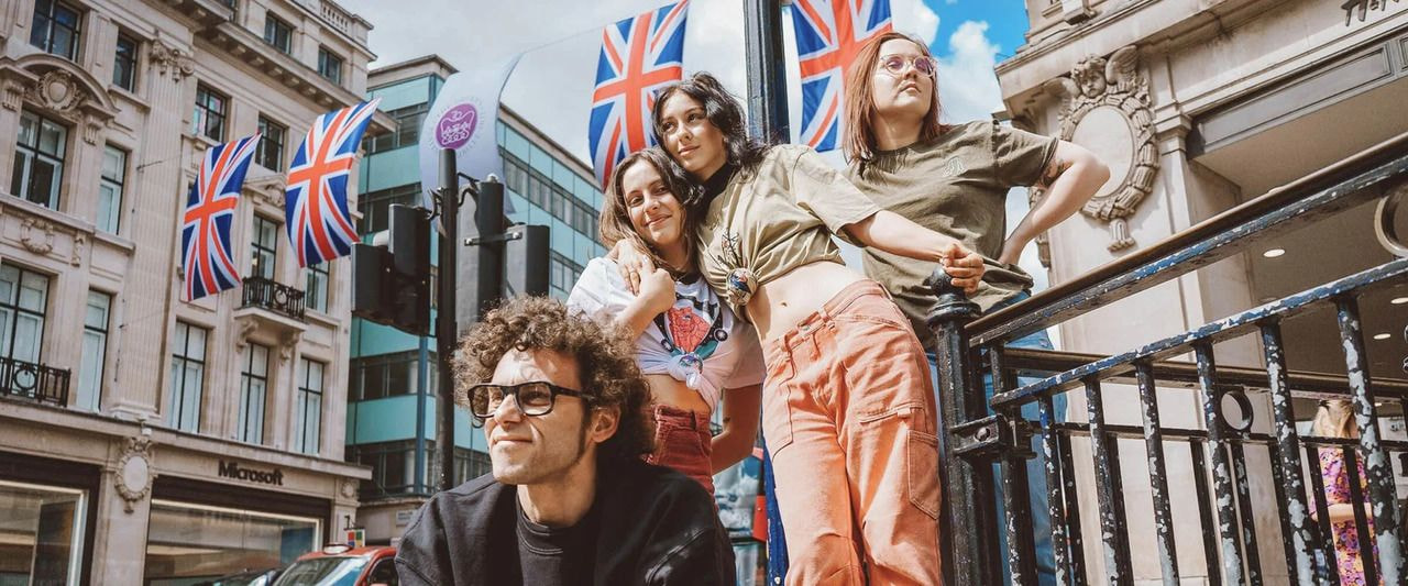Group of four young adults posing on a city street lined with Union Jack flags, wearing casual custom apparel in front of historic buildings on a sunny day.