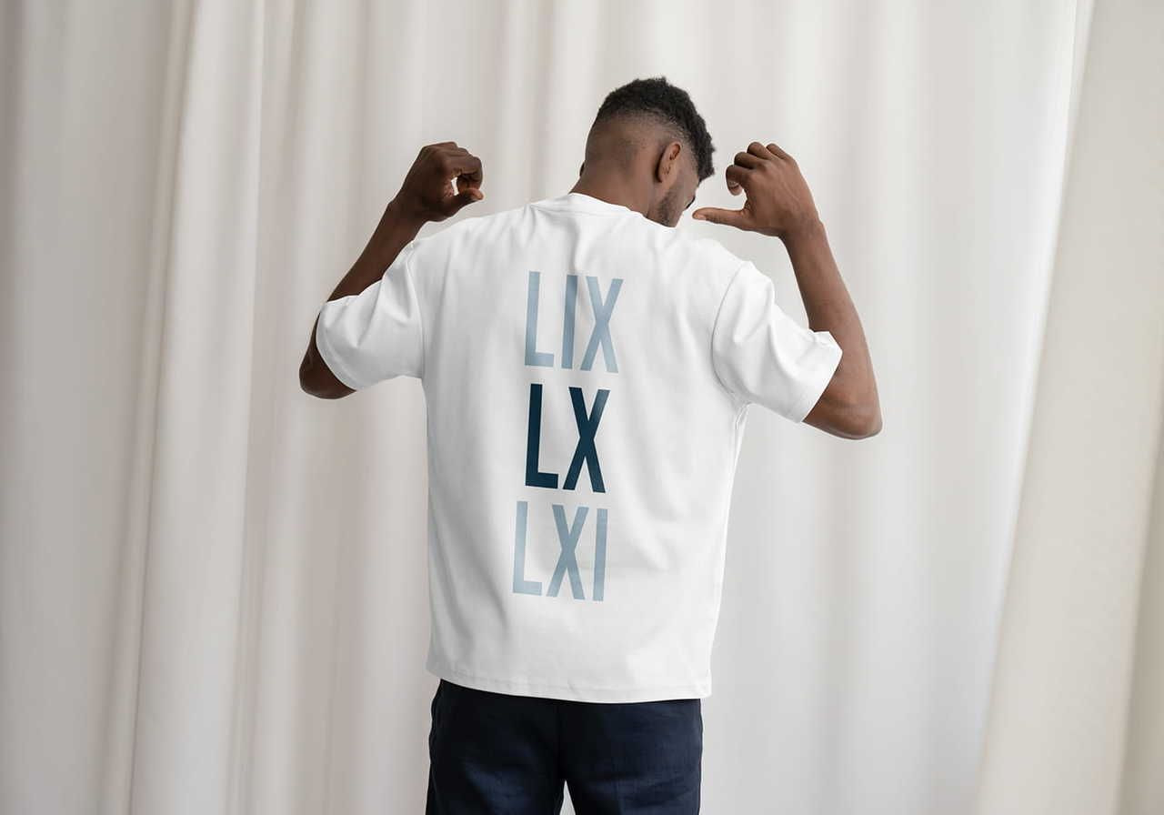 A man facing away, flexing arms, wears a white t-shirt with blue Roman numerals LIX, LX, LXI. The background is soft white curtains.