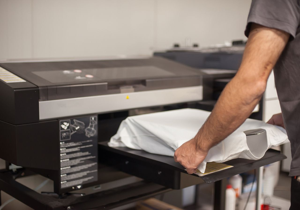 A worker places a white t-shirt onto the platen of a direct-to-garment printer in a print shop.