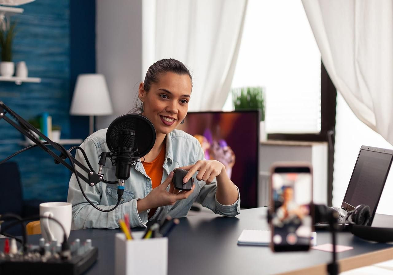 A woman smiles while recording a product presentation using a microphone and a phone on a tripod.