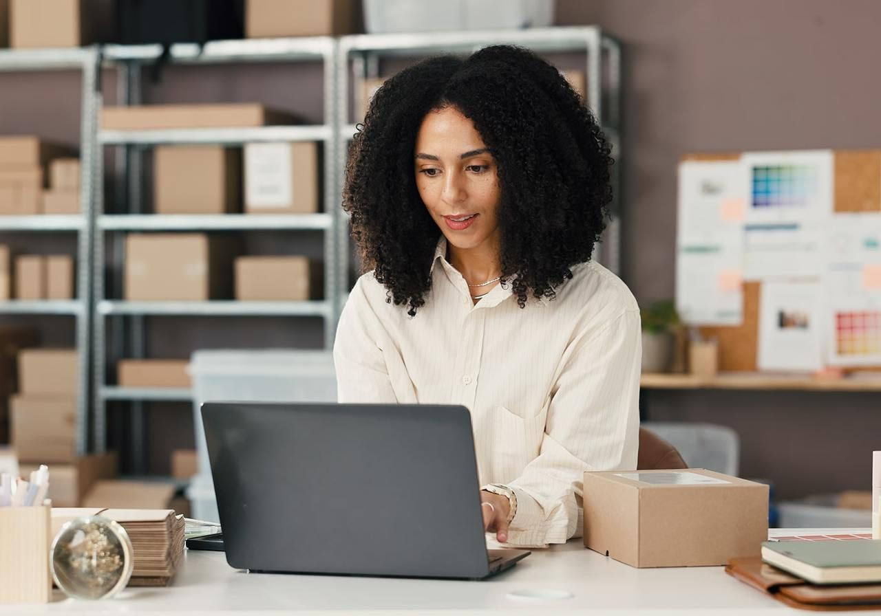 A woman with curly hair works on a laptop, searching for Etsy alternatives, in a home office surrounded by packages and stationery.