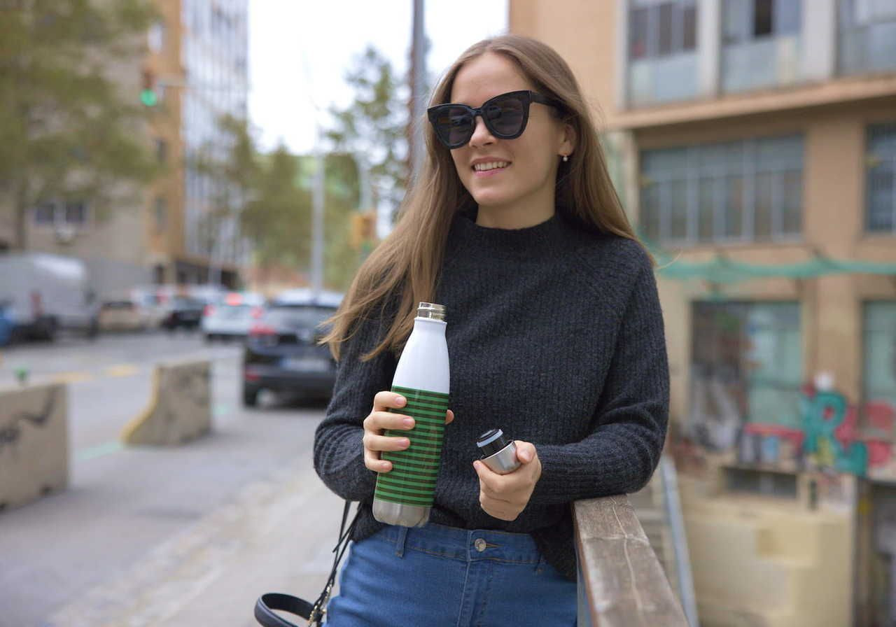 A woman with long brown hair, wearing sunglasses and a black sweater, is standing near a railing, holding a water bottle and a small container.