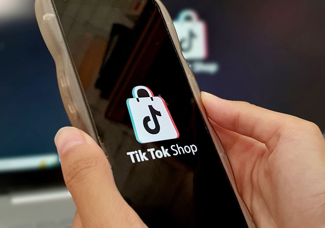 A hand holds a smartphone displaying the TikTok Shop logo, with the same logo blurred on a screen in the background.