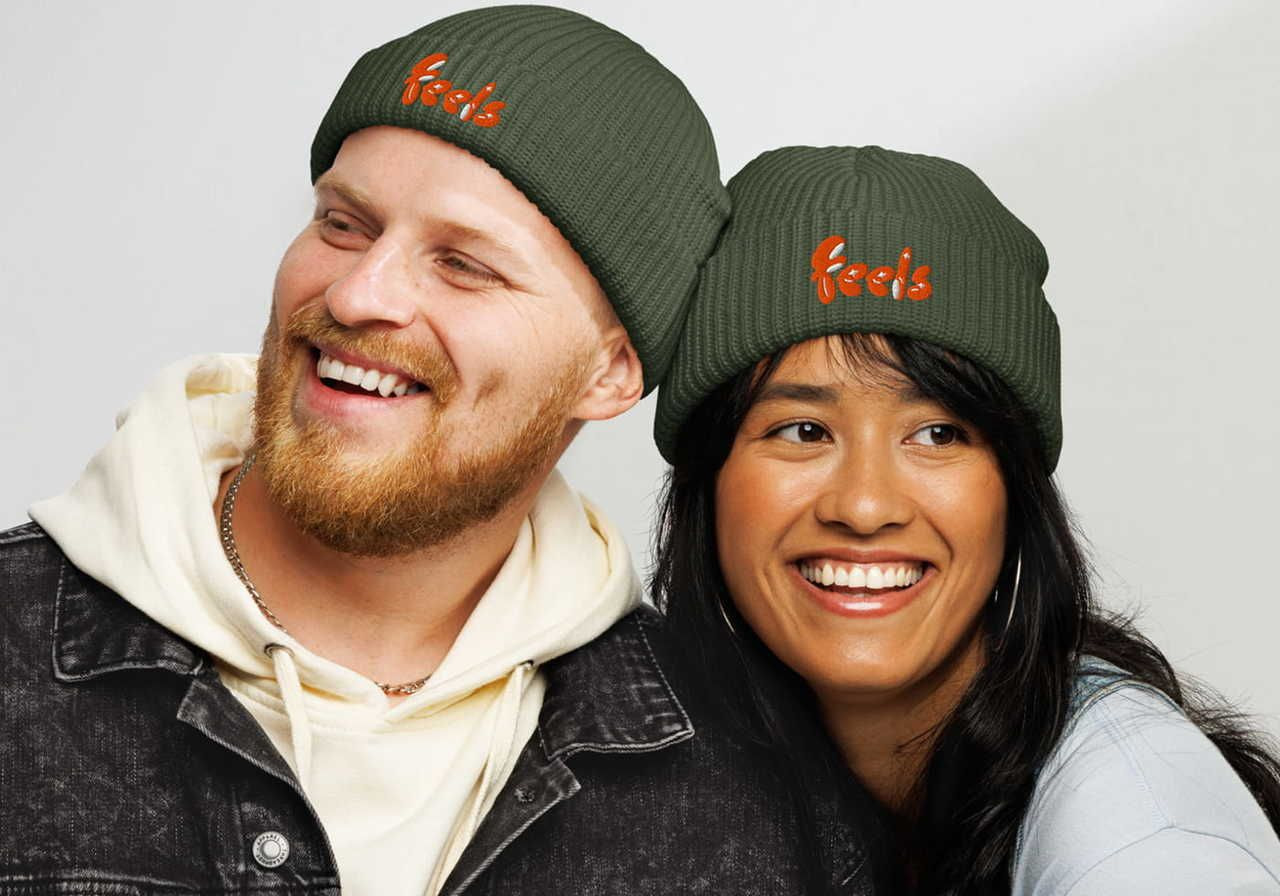 A smiling man and woman wearing green beanies with "feels" embroidered in orange.
