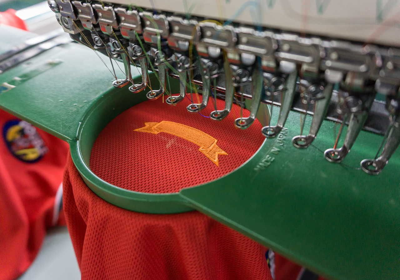 Close-up of a sewing machine embroidering an orange ribbon on red t-shirt, framed by a green hoop.