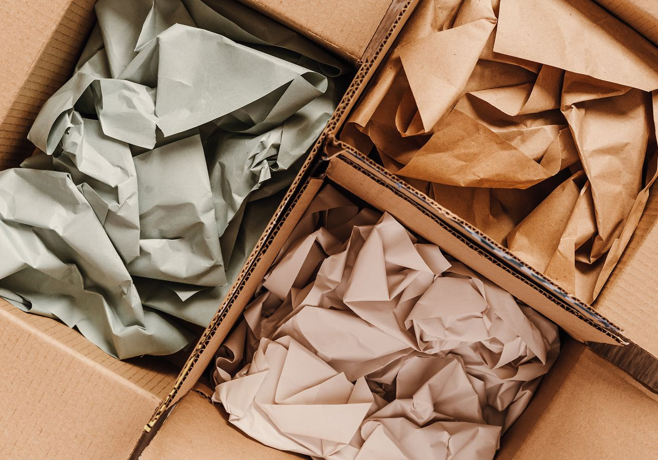 Three cardboard boxes filled with crumpled sage green, kraft brown, and blush pink paper show eco-friendly packaging materials.