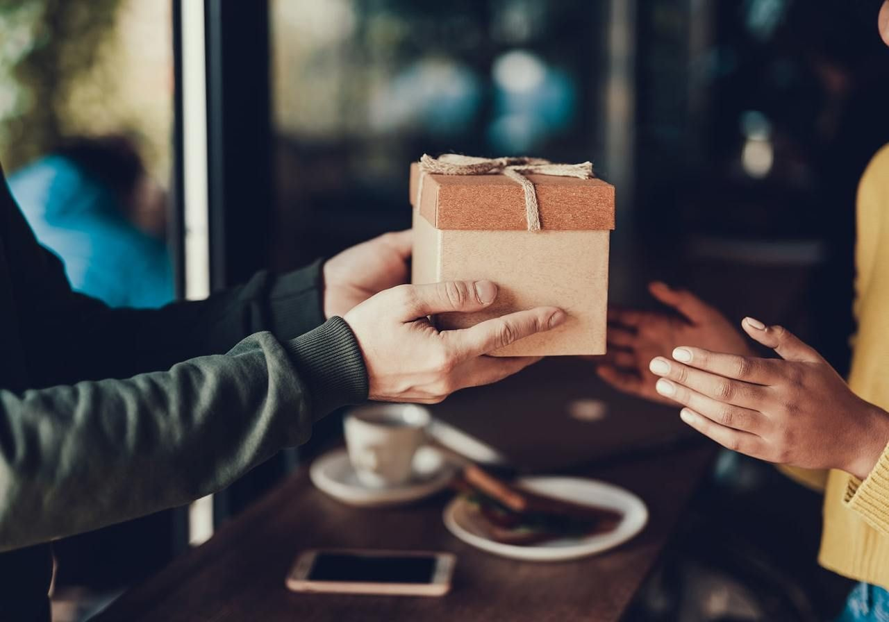 A man presents a wrapped gift to a smiling woman in a warm, inviting setting.