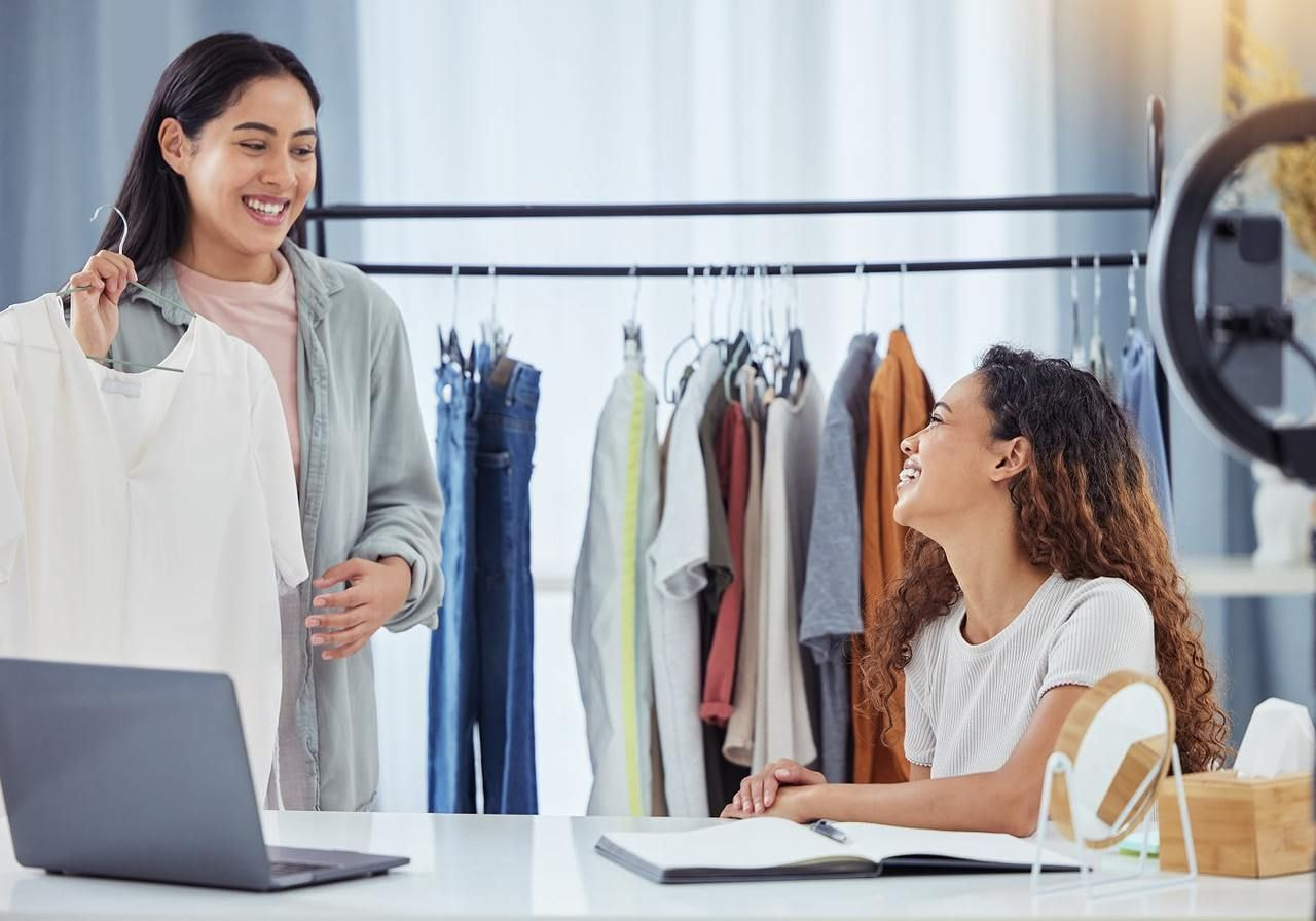 Two women are in a room; one holds a white shirt, smiling, while the other sits, laughing. Behind them, clothes hang on a rack.