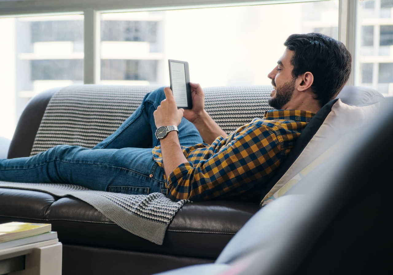 A man lying on a couch, reading an e-reader, with a relaxed smile on his face, wearing a plaid shirt and jeans.