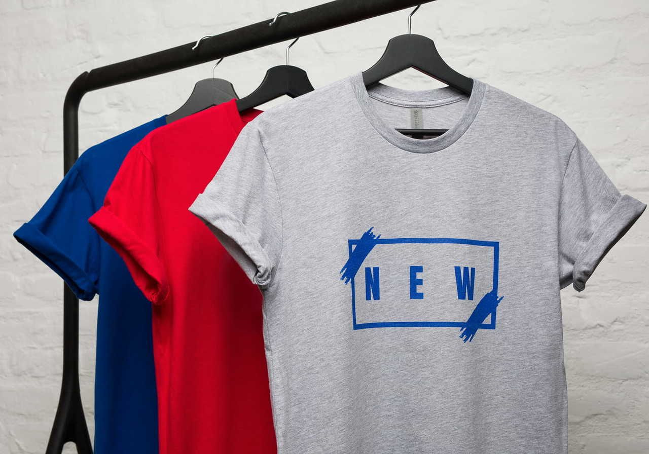 Three t-shirts hanging on a rack: one gray with "NEW" printed on it, and two in the background, one blue and one red.