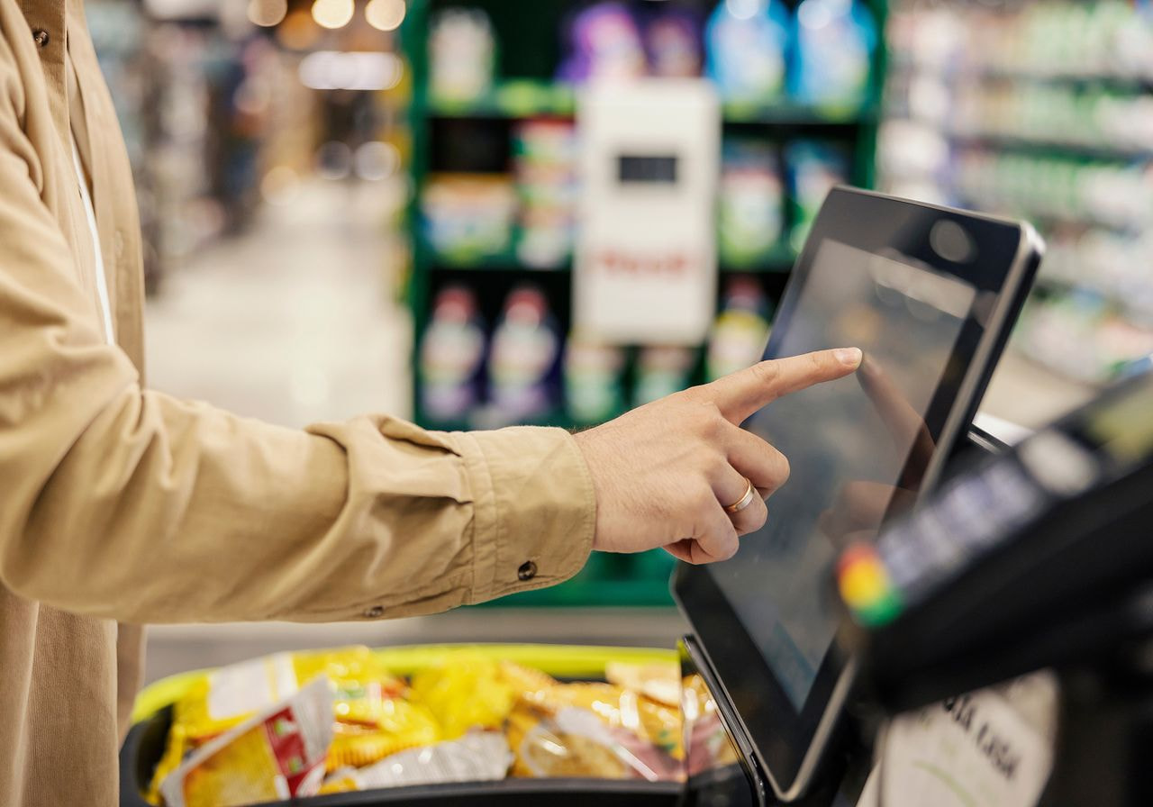 A shopper taps the touchscreen of a self-checkout kiosk in a grocery store with a cart of snacks nearby.