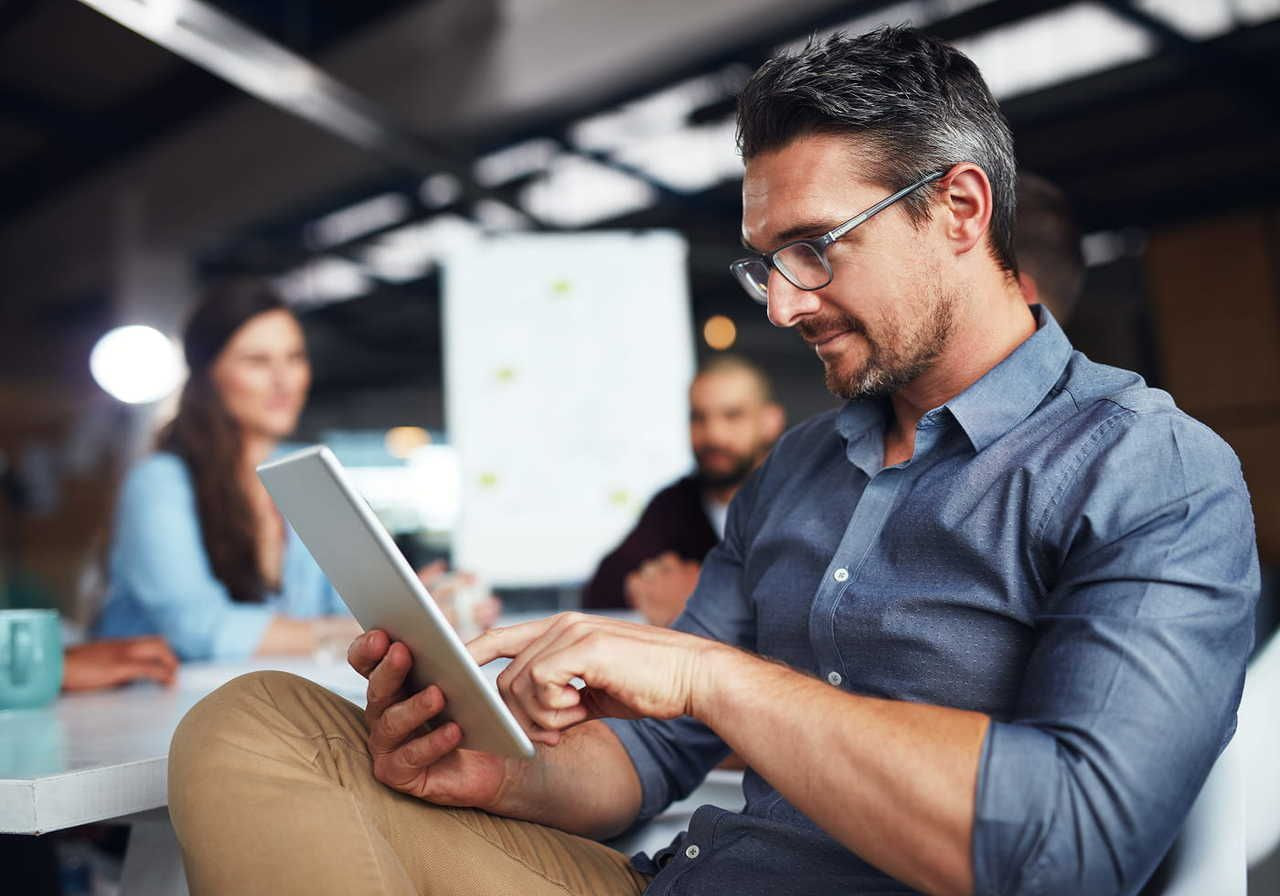 A man using a tablet to find types of shirt printing in a modern office setting, surrounded by colleagues in a meeting.