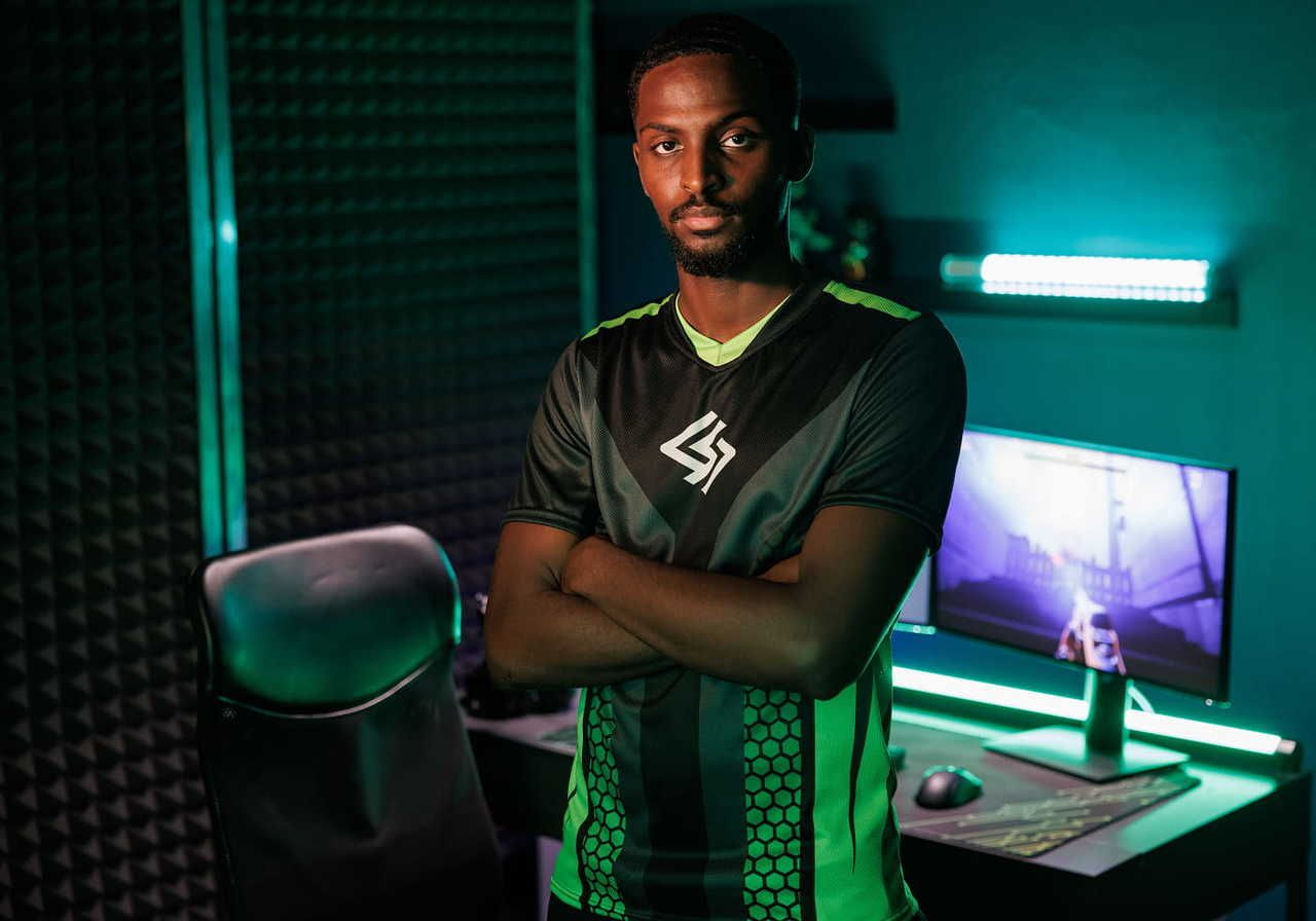 A man in a green and black esports jersey stands confidently with arms crossed, with a gaming setup behind him.