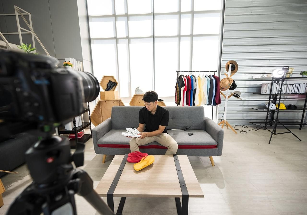 A man sits on a gray sofa in a bright room, presenting custom products and filming them using a camera for YouTube.