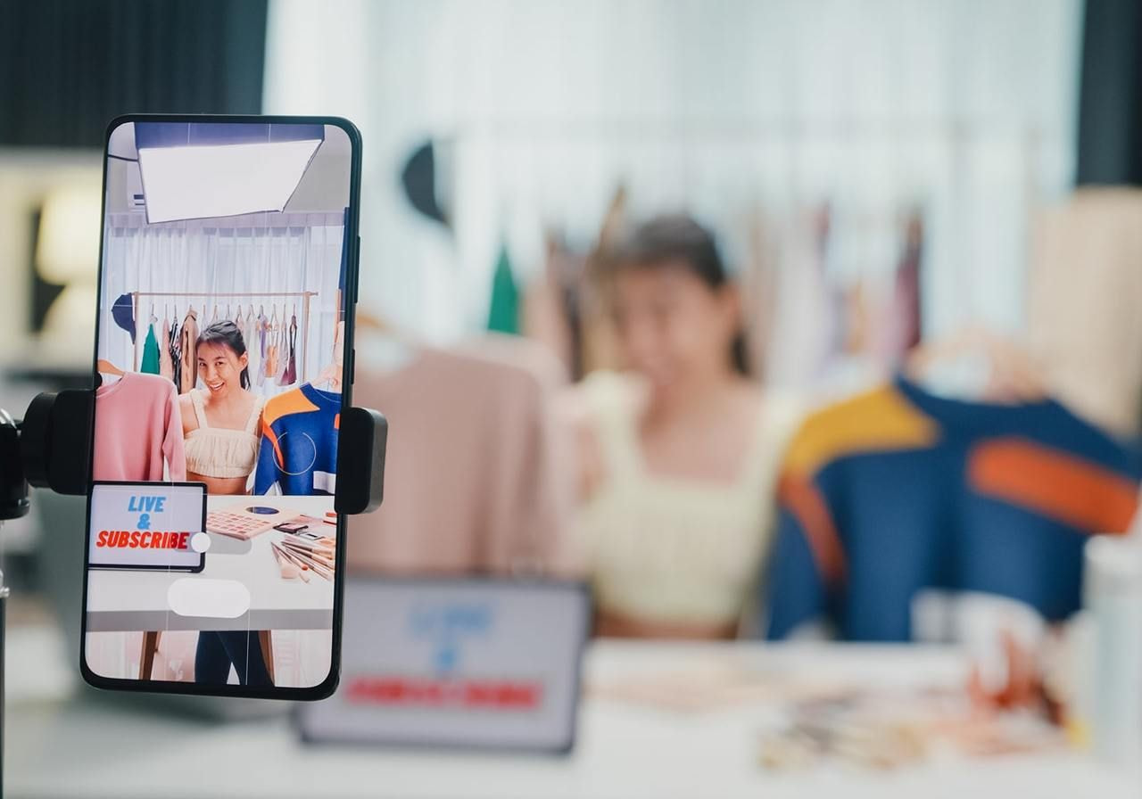 A woman livestreams fashion content featuring sweaters displayed on a smartphone. A “Live & Subscribe” sign is prominent.