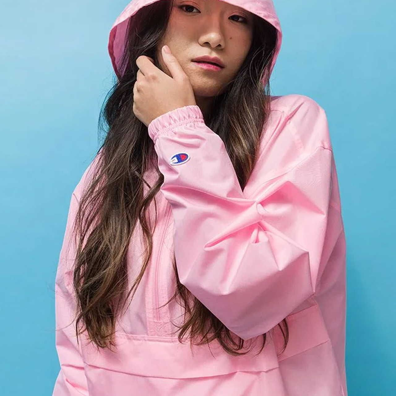 Young woman in a pink hoodie against a blue background, with one hand touching her face. The hoodie has a small embroidered logo on the sleeve.